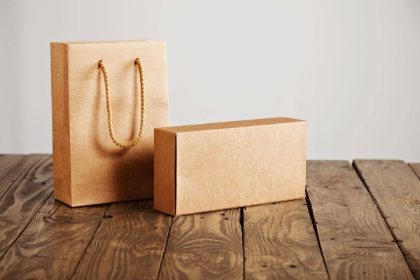 Brown kraft paper shopping bag with rope handles alongside a plain kraft cardboard box on a rustic wooden surface.